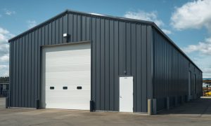 Custom 30x80 metal building with black steel siding, a tall white overhead door, side entry, and bollards for protection