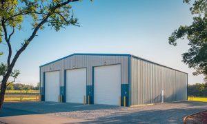 45x50 commercial steel building auto shop with three large doors, blue and gray PBR panels, and a gravel lot surrounded by trees