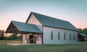 Small steel church building with seating for 100, arched windows, red trim, and a covered front canopy on open grass
