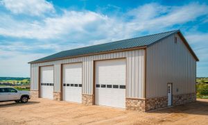 Three car steel building garage with stone base, in white and beige, with stone wainscot and side entry door on a rural lot