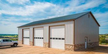 Three car steel building garage with stone base, in white and beige, with stone wainscot and side entry door on a rural lot