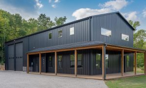 Two-story black metal building shop house with wraparound wood porch, large garage bay, and upper windows in a wooded setting