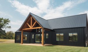 Black steel building home with large timber porch, 2 bedrooms, steep pitched roof, and tall windows on a grassy open property