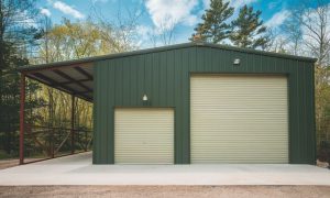 A dark green metal building used as a garage with two roll-up doors and a red-framed side lean-to canopy on a forested country property in Denver Colorado