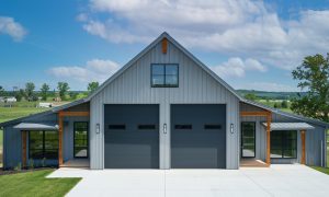 One-story barndominium metal building with RV garage, 3 bedrooms, twin black doors, and wood-trimmed wings on a concrete pad in rural Texas